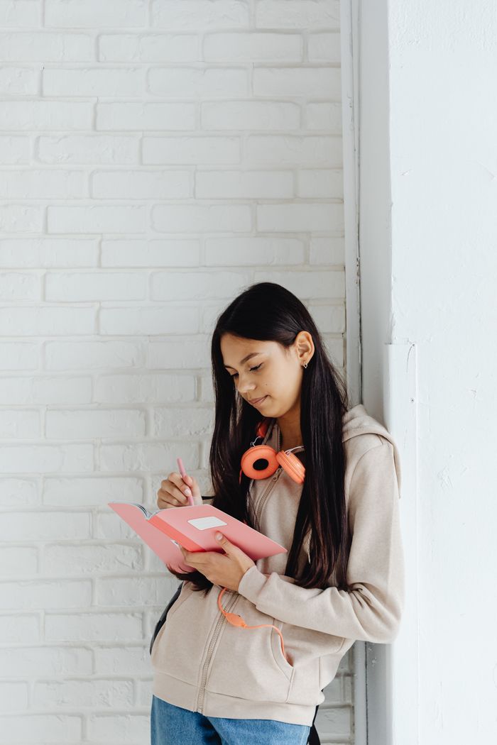 Teen girl writing in a notebook, leaning against a white brick wall, with headphones around her neck.